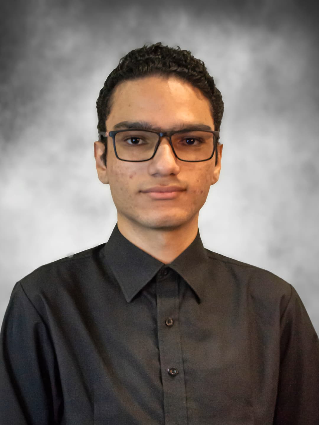 Foto minha, um retrato de busto onde estou vestindo uma camisa social preta, tenho cabelos escuros e cacheados, e uso óculos de grau com armação preta. O fundo da foto é um cinza com textura sutil.